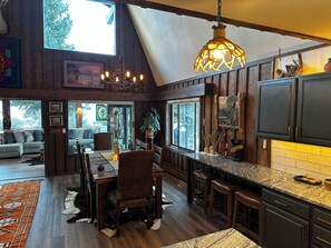 Un intérieur rustique comprenant un coin repas, une cuisine et un espace de vie, avec des murs en bois, un lustre et de grandes fenêtres.