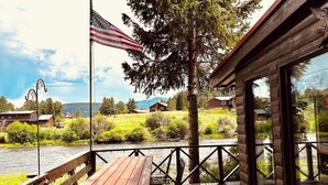 Eine Holzterrasse mit Picknicktisch und Stühlen, mit Blick auf einen Fluss und ein bewaldetes Gebiet, im Hintergrund sind Häuser zu sehen.