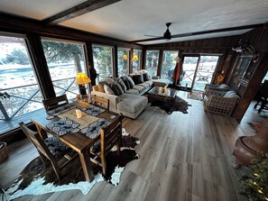 Ein geräumiger Wohnbereich mit Holzboden, einer Sofagarnitur, einem Esstisch mit gemusterten Untersetzern und großen Fenstern mit Blick ins Freie.