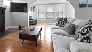 A living room with a fireplace, a flat-screen TV mounted on the wall, a grey sofa with patterned cushions, and a wooden coffee table.
