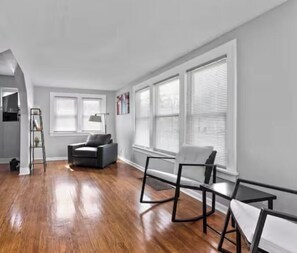 A spacious room with hardwood flooring, a grey sofa, a white armchair, and a small table. - 3 Bedrooms 2 baths 10 minutes from SU, Downtown, Hospitals and Airport (Syracuse)