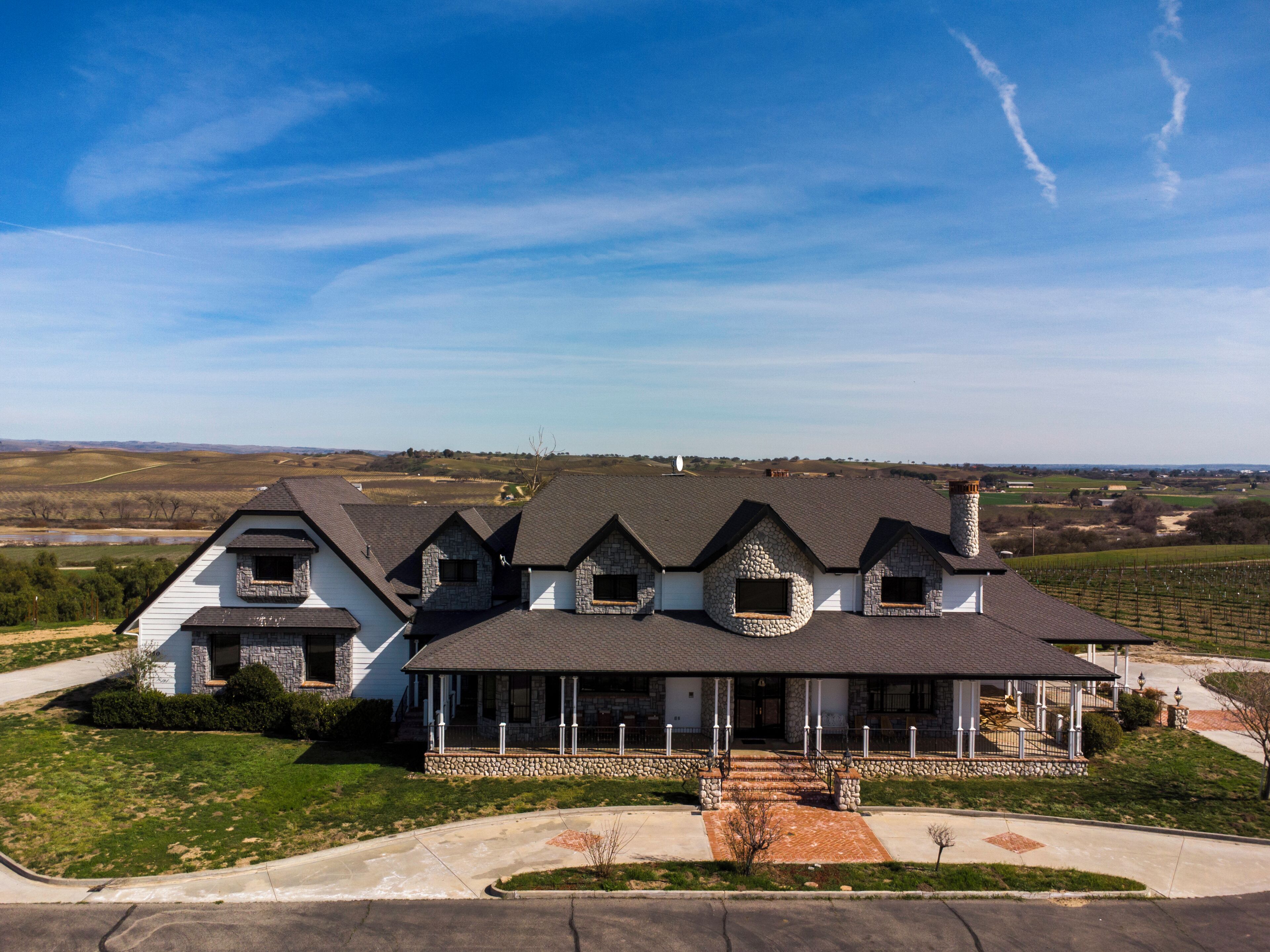 Ein großzügiges, zweistöckiges Haus mit Naturstein-Fassade und mehreren Giebeln, umgeben von einem gepflegten Rasen und einem Weinberg im Hintergrund.