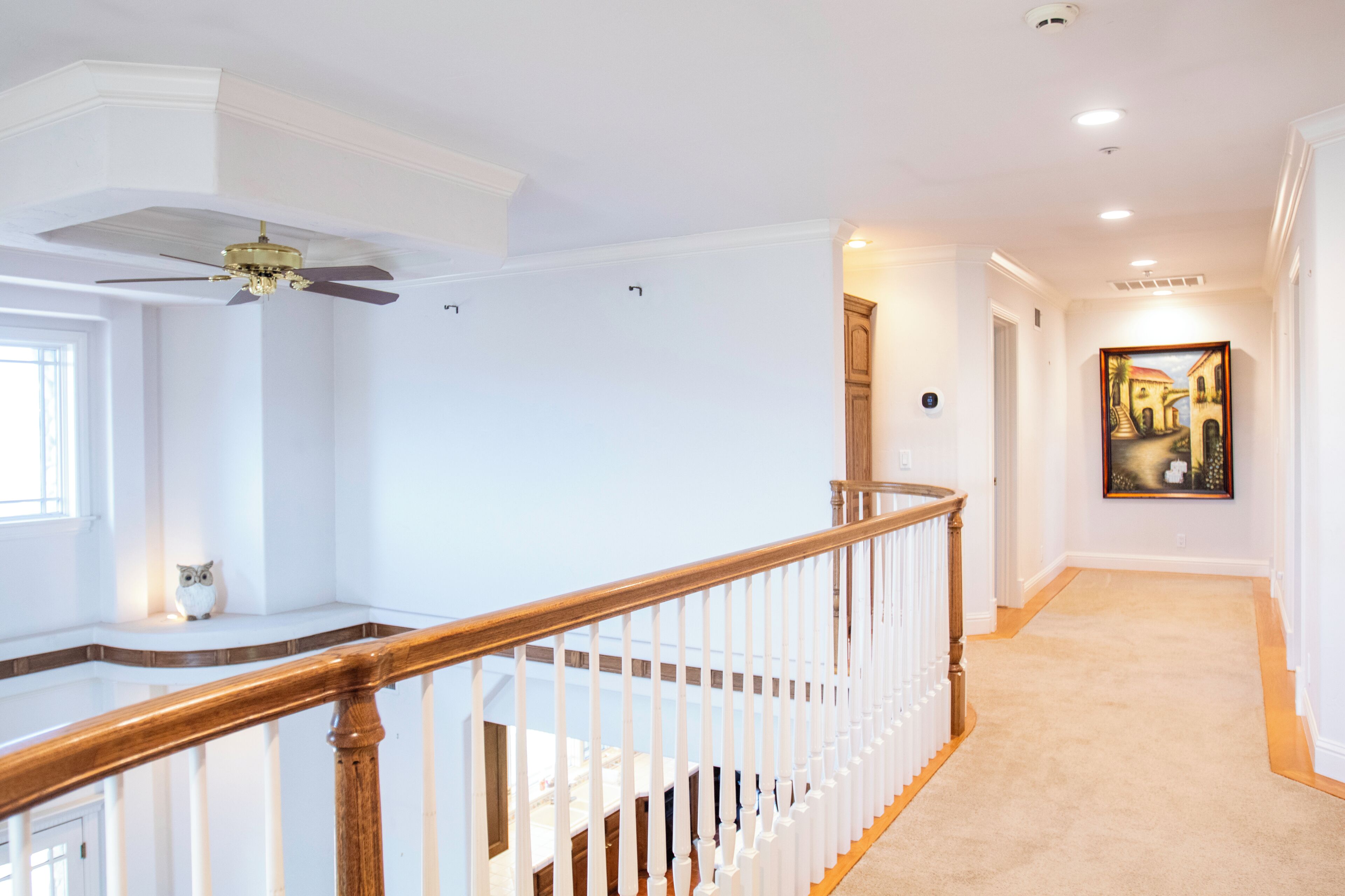Un escalier avec une rampe en bois menant à l’étage supérieur, un ventilateur de plafond et un couloir orné d’un tableau.