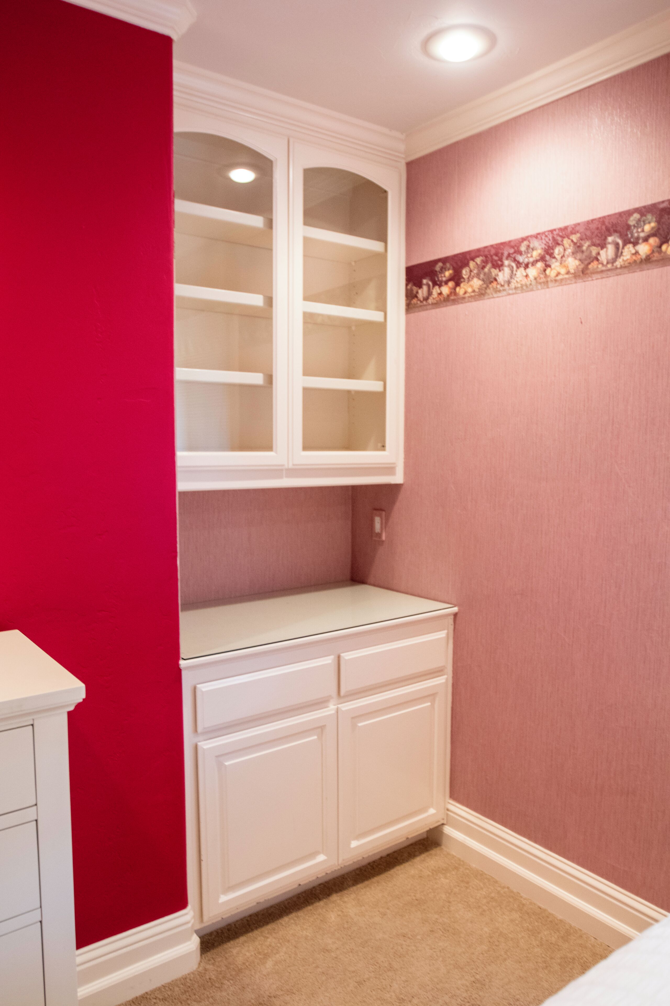 A room with a built-in white shelving unit against a red wall, a beige carpet, and a decorative border on the wall.