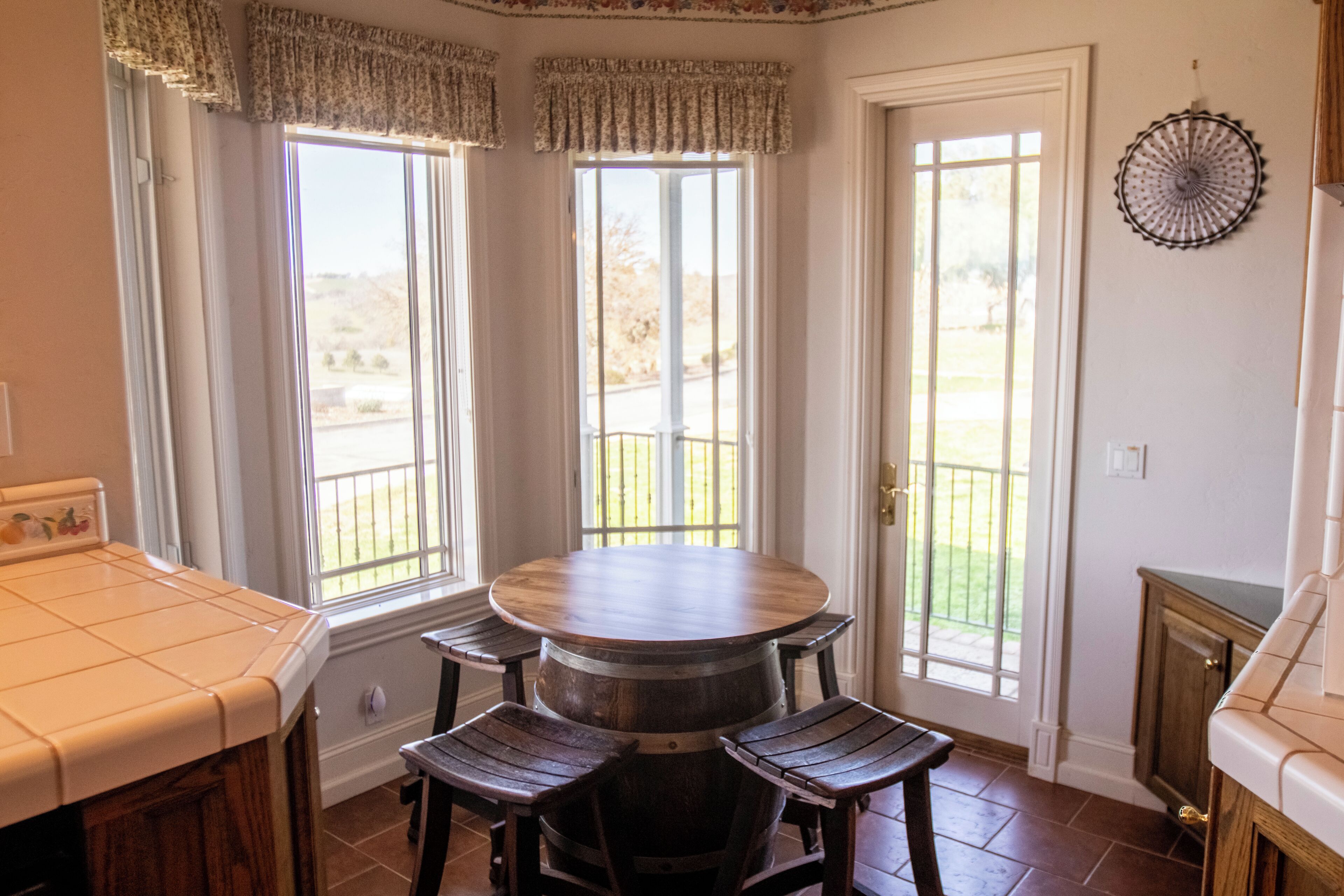 A dining area with a round table and four wooden chairs, large windows with curtains, and a view of a garden.