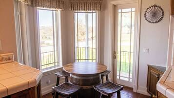 A dining area with a round table and four wooden chairs, large windows with curtains, and a view of a garden.