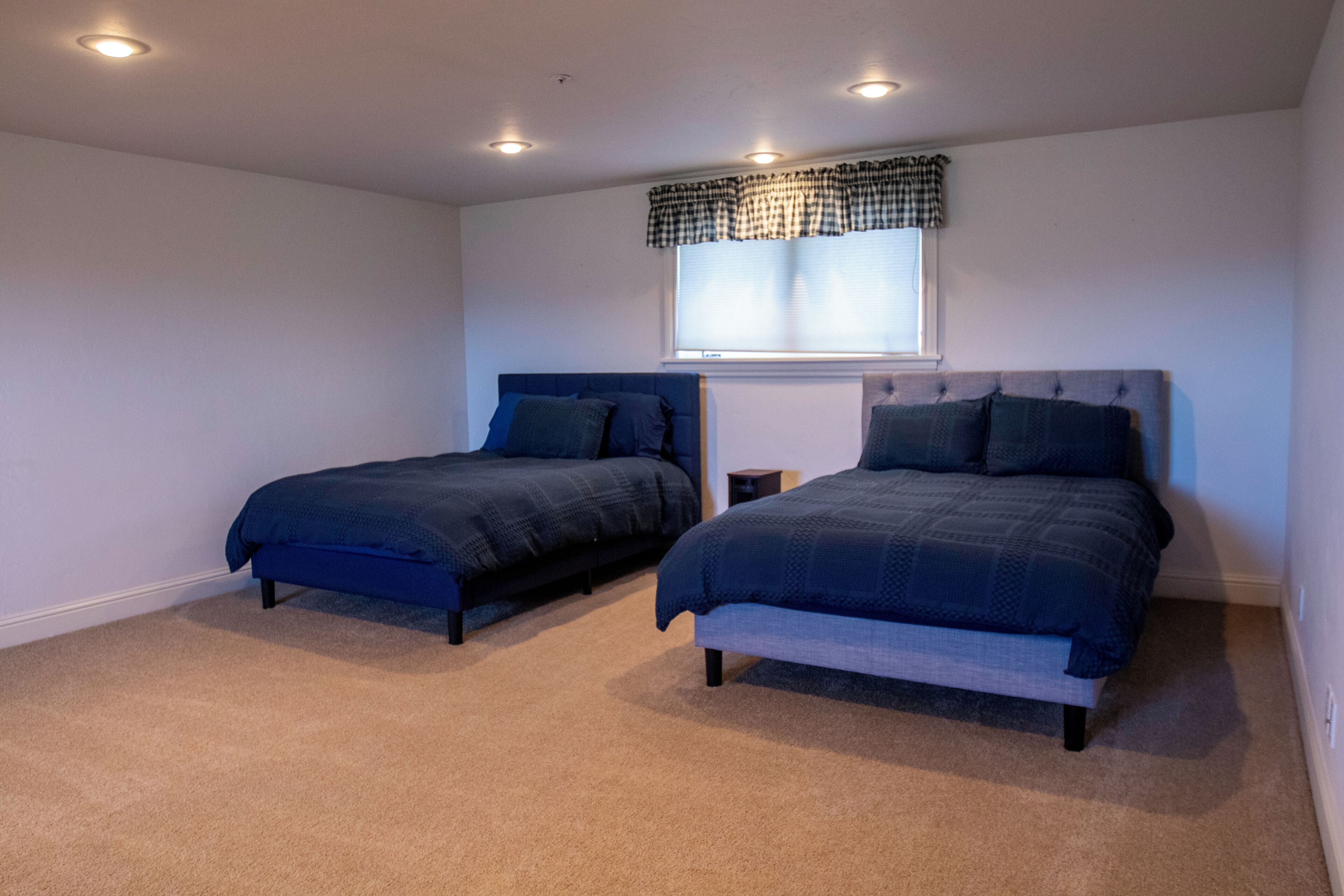 A bedroom with two beds, a window with blinds, and recessed ceiling lights.