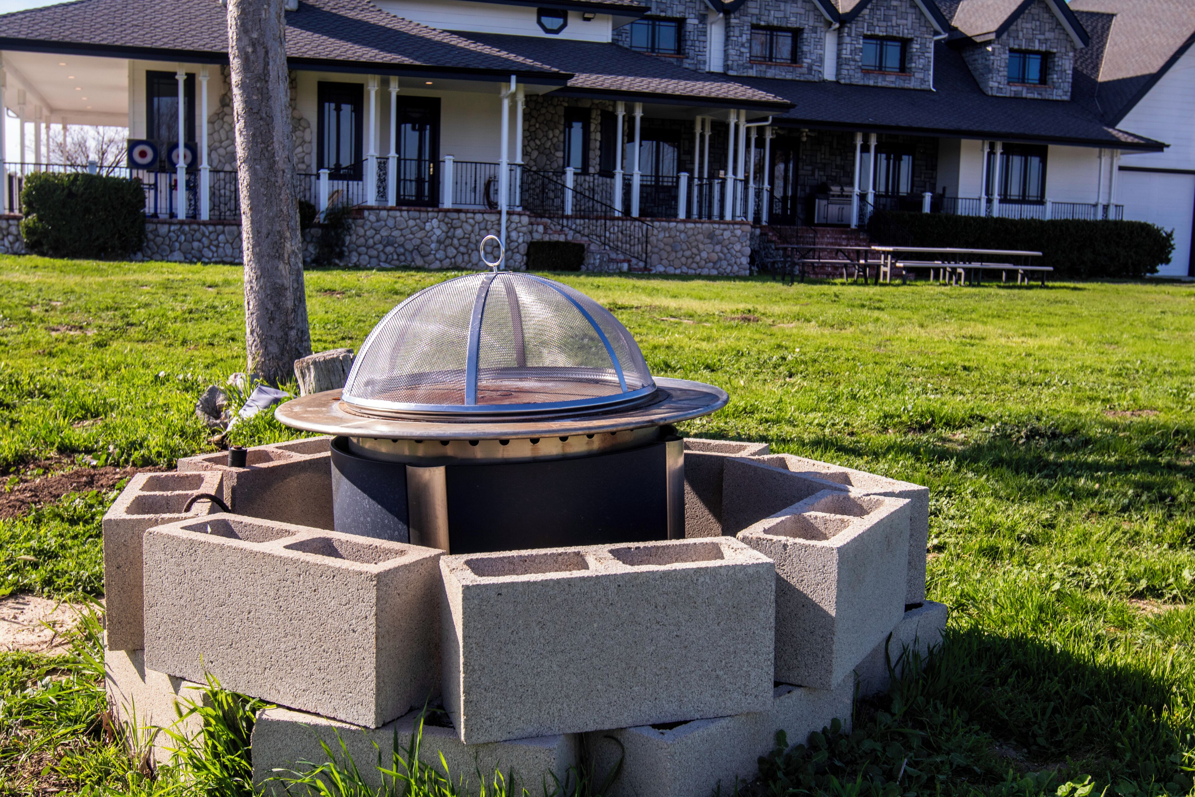 A fire pit with a protective cover, surrounded by concrete blocks, in a grassy area with a building in the background.
