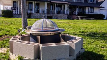 A fire pit with a protective cover, surrounded by concrete blocks, in a grassy area with a building in the background.