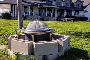 A fire pit with a protective cover, surrounded by concrete blocks, in a grassy area with a building in the background. - Santellan Manor (Paso Robles)