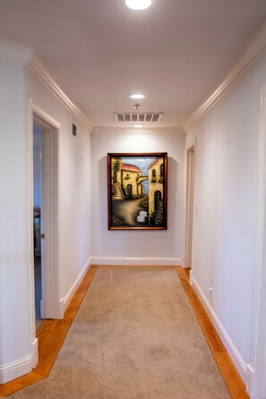 A hallway with a painting on the wall, carpeted floor, and white walls. - Santellan Manor (Paso Robles)