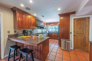 Una cocina con armarios de madera, una isla central con taburetes, un cubo de basura de acero inoxidable y vistas al exterior.