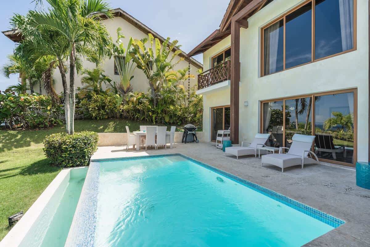 Ein Pool mit Liegestühlen und einer Terrassenfläche, umgeben von üppiger Bepflanzung und einem zweistöckigen Gebäude mit großen Fenstern.