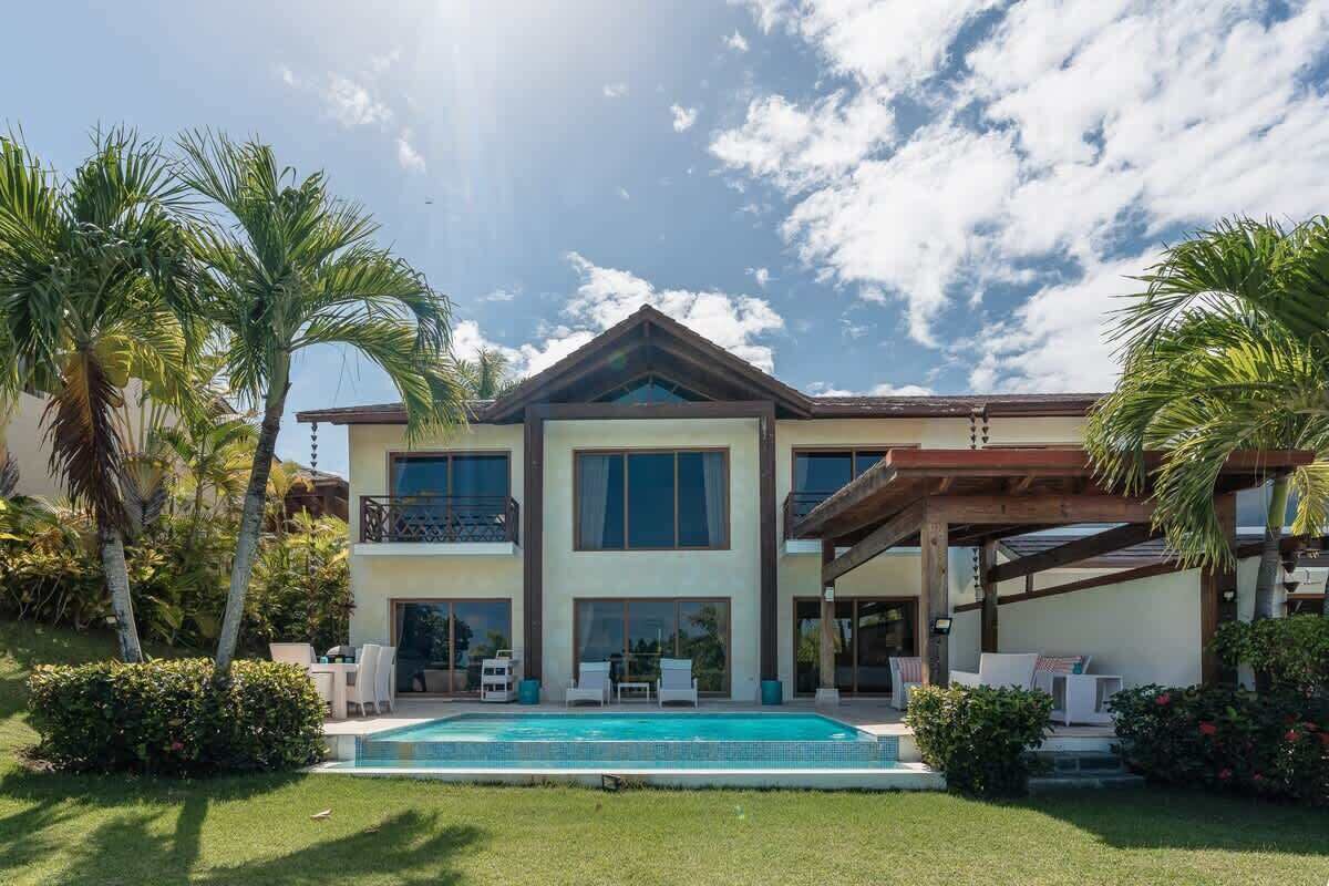 Una casa de dos pisos con piscina, rodeada de palmeras y un césped cuidado.