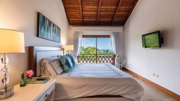 Ein Schlafzimmer mit Holzboden, einem Bett mit weißer Bettwäsche, einem Nachttisch mit Lampe, einem Fernseher an der Wand und einem Fenster mit Blick ins Grüne.
