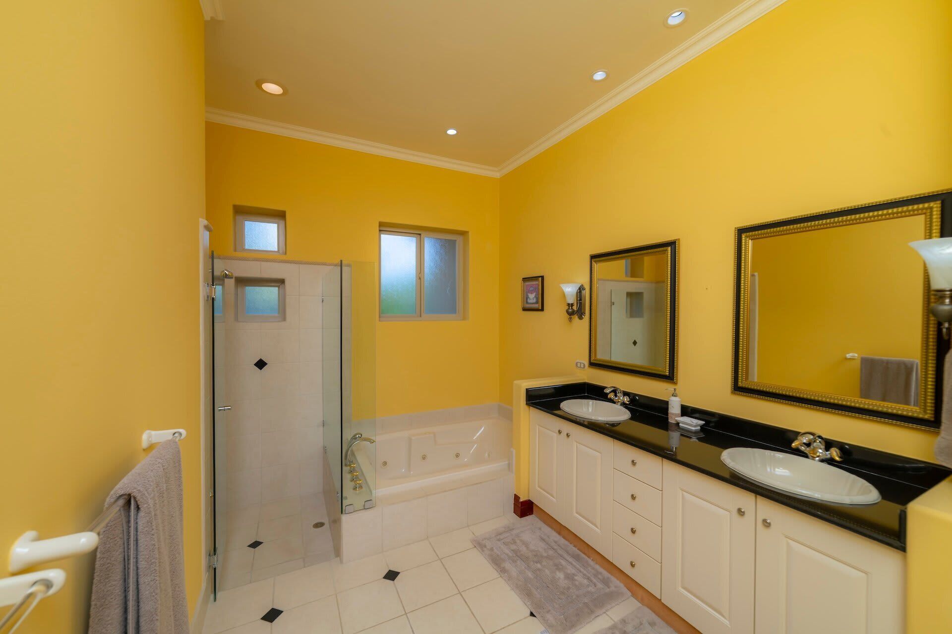 Baño con paredes amarillas, cabina de ducha de vidrio, bañera, dos lavamanos y un espejo grande.