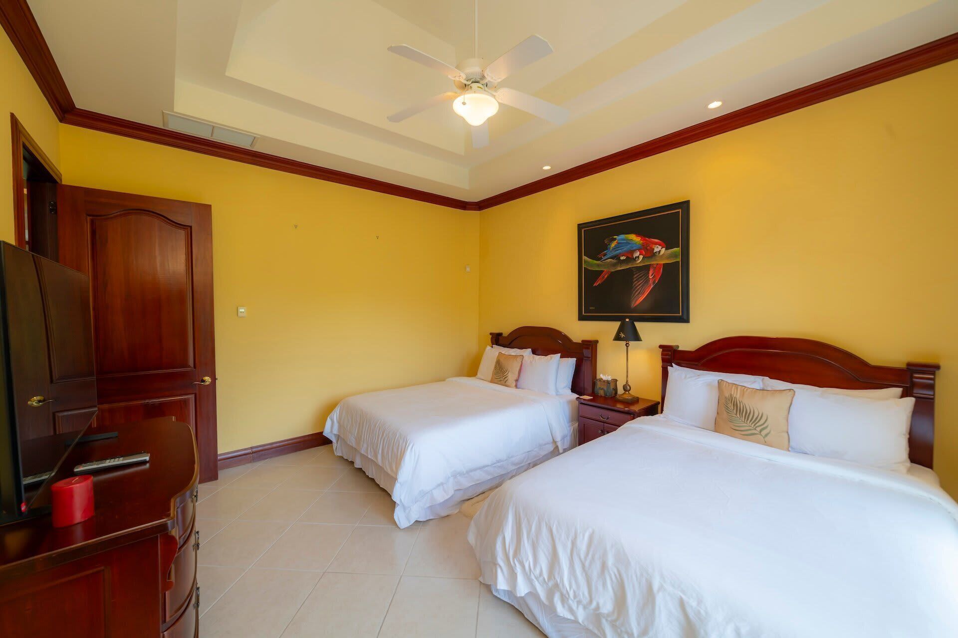 Habitación de hotel con dos camas, un ventilador de techo, un cuadro de un loro y un televisor sobre un mueble de madera.