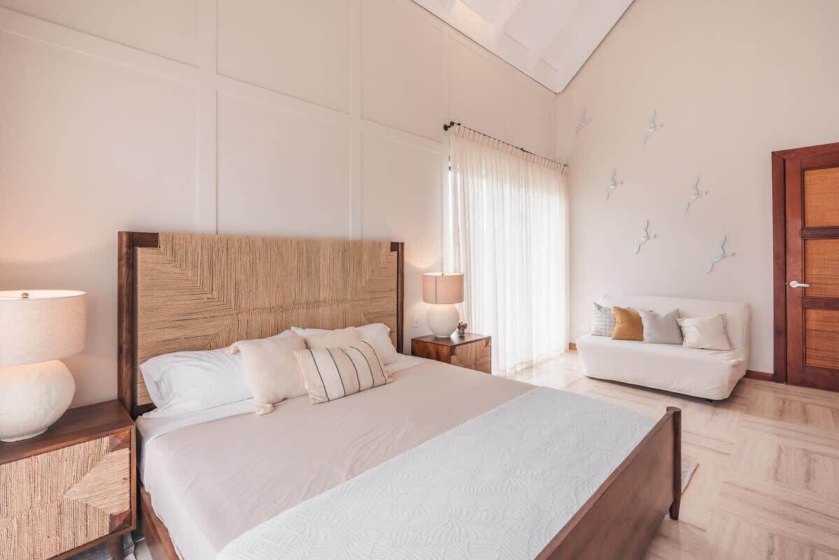 A neatly arranged bedroom with a large bed, bedside tables, a sofa, and a window with curtains.