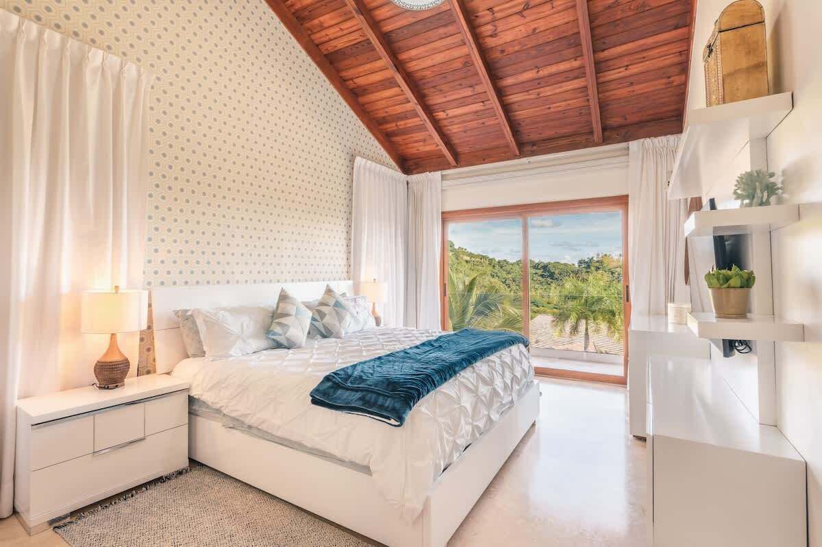 A bedroom with a bed, nightstands, a lamp, a window with a view, and a ceiling with wooden beams.