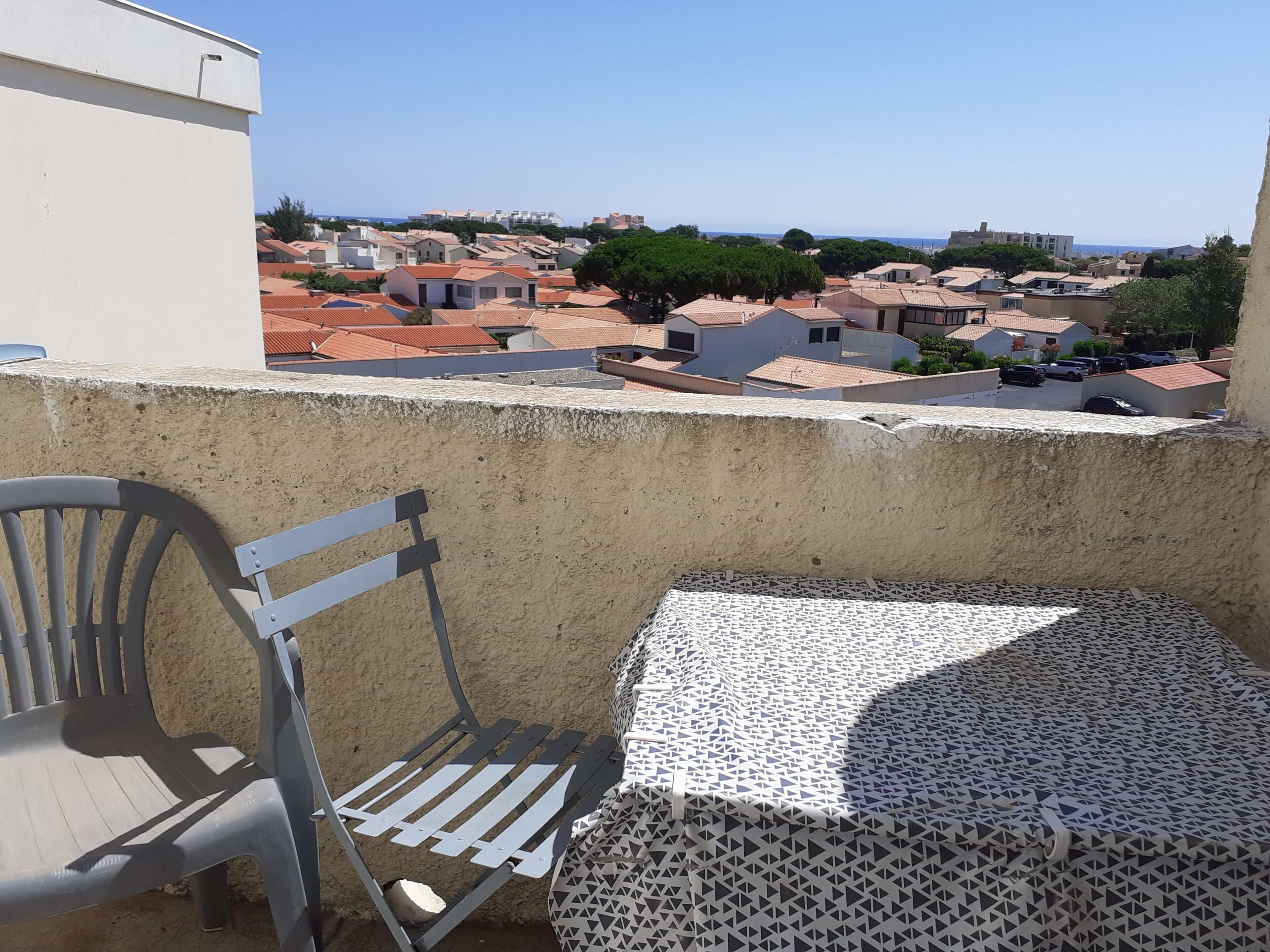 Un balcón con una silla de metal y una mesa, con vistas a una zona residencial con casas y un cielo despejado.