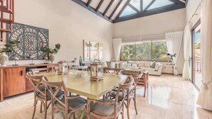 Uma sala de estar ampla com teto alto, uma mesa de jantar com cadeiras, um sofá e uma janela grande com cortinas.