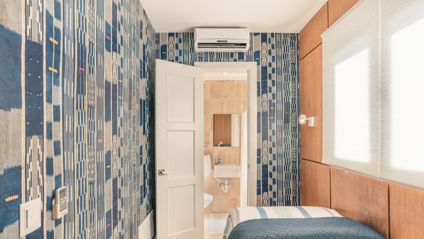 A bedroom with a bed, an air conditioning unit, a bathroom with a sink and mirror, and patterned wallpaper. - Apt with 2BR & balcony beach access in Portillo (Las Terrenas)
