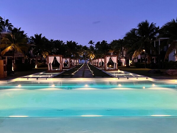 A pool with a clear view of the water, surrounded by palm trees and a pathway leading to a building.