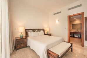 A bedroom with a large bed, two bedside tables with lamps, a wooden bench, a bathroom with a sink and mirror, and a door leading to another room.
