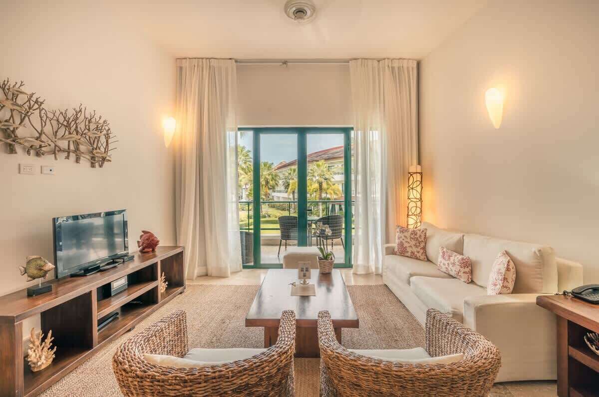 Sala de estar con televisor, sofá y sillas de mimbre. Cuenta con un ventanal con cortinas y vistas a palmeras.