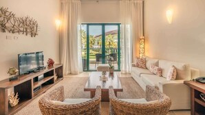 Un salon avec un téléviseur, un canapé et des fauteuils en osier. Il possède une grande fenêtre avec des rideaux et offre une vue sur des palmiers.