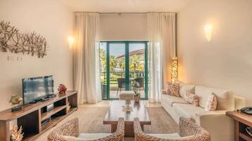 Sala de estar con televisor, sofá y sillas de mimbre. Cuenta con un ventanal con cortinas y vistas a palmeras.