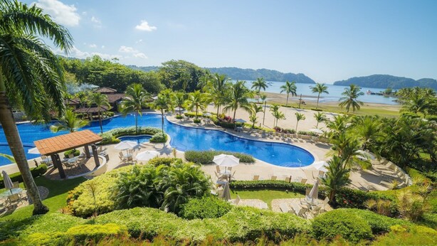 Un complexe hôtelier doté d’une grande piscine sinueuse, entourée de palmiers et donnant sur une plage.