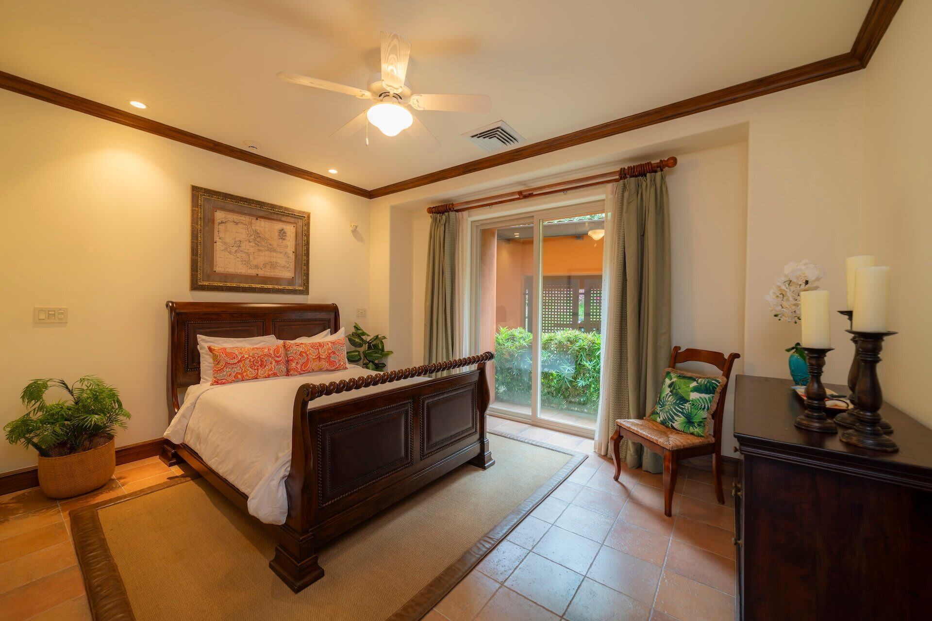 A bedroom with a wooden bed, a ceiling fan, a large window with curtains, and a view of greenery outside.