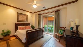 A bedroom with a wooden bed, a ceiling fan, a large window with curtains, and a view of greenery outside.