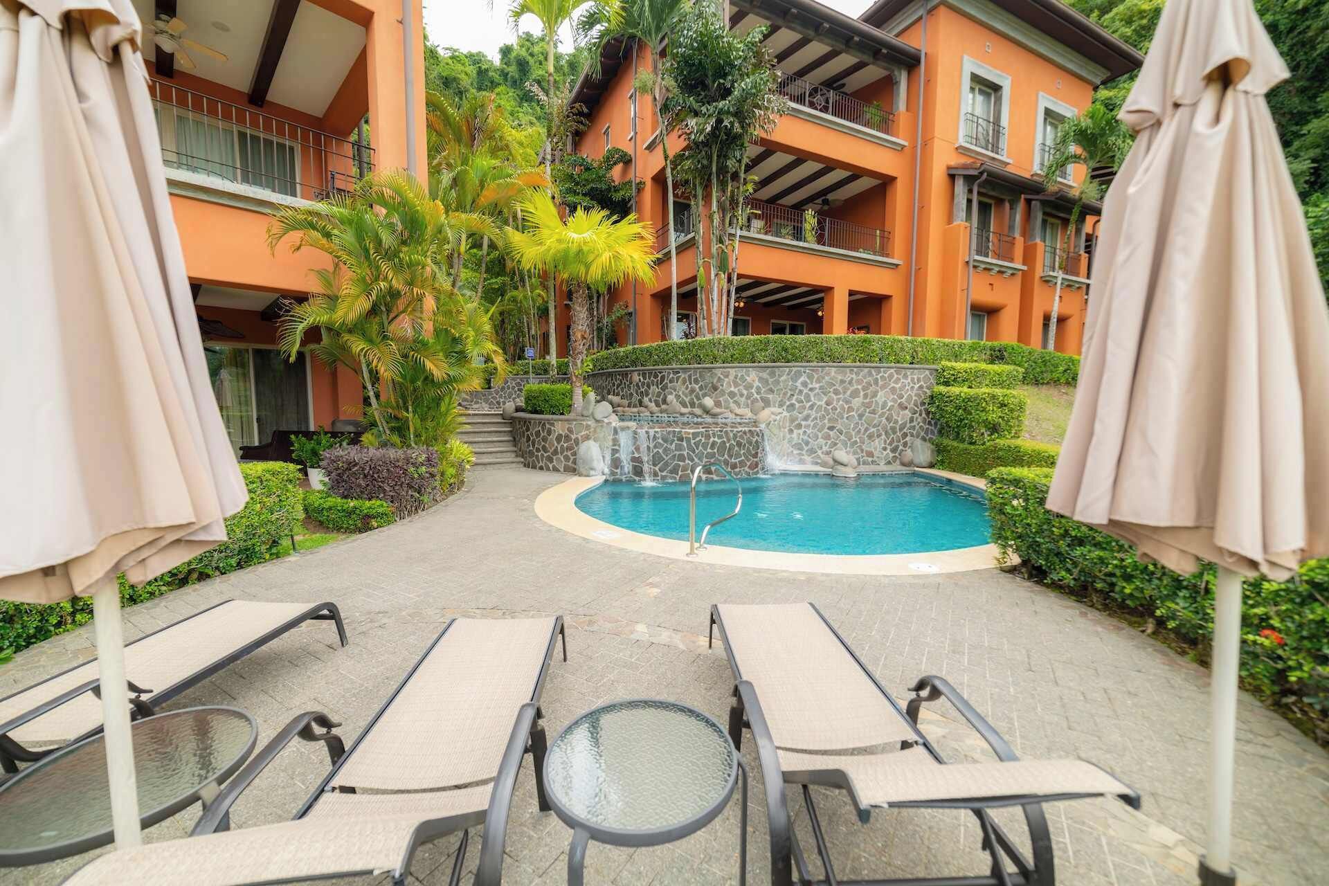 A pool area with lounge chairs and umbrellas, surrounded by a building with balconies and greenery.
