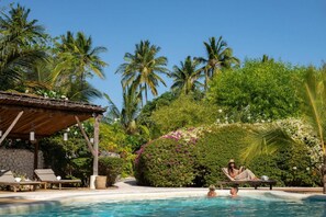 Ein Poolbereich mit Liegestühlen, einer überdachten Sitzgelegenheit und üppiger tropischer Vegetation.