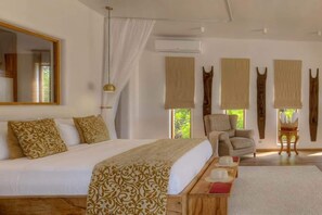 A bedroom with a large bed, a chair, a small table with hats, and a window with blinds. - Indian Ocean Beachfront Sanctuary (Paje)