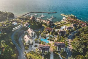 Eine Vogelperspektive zeigt einen Urlaubsort mit mehreren Gebäuden, einem zentralen Schwimmbecken und einer Marina mit Booten.