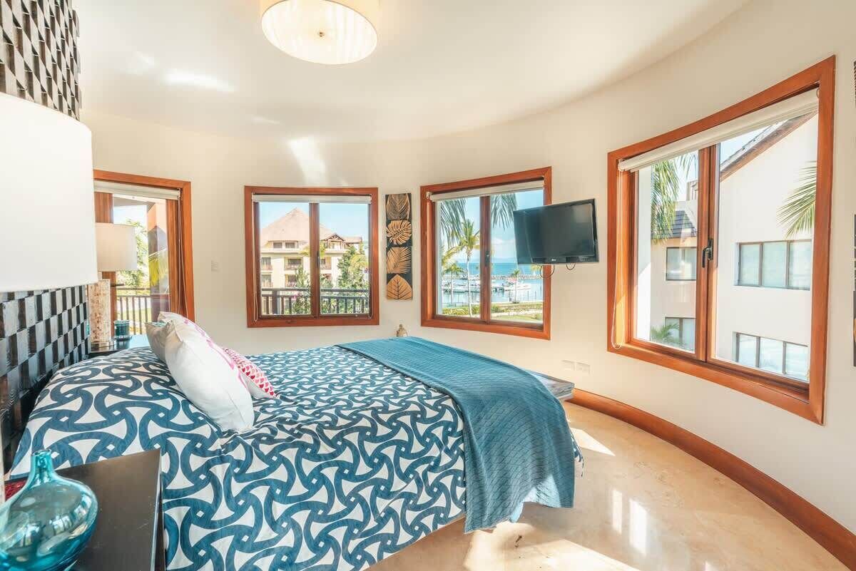 Une chambre à coucher avec un lit, une télévision et de grandes fenêtres donnant sur une plage et des immeubles.
