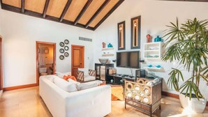 Uma sala de estar espaçosa com um sofá branco, uma mesa de centro de madeira e uma TV de tela plana. Há relógios decorativos na parede e prateleiras com diversos objetos.