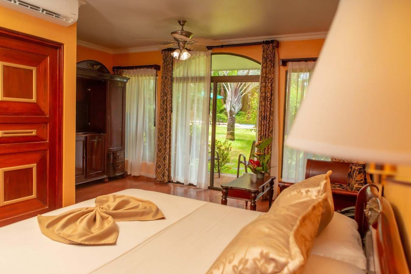 A hotel room with a bed, curtains, a ceiling fan, and a view of a garden.