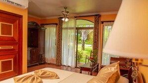 A hotel room with a bed, curtains, a ceiling fan, and a view of a garden.