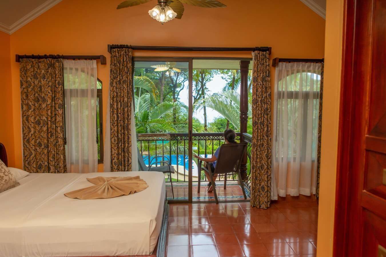 Una habitación de hotel con cama, balcón y vistas a una piscina, además de ventilador de techo.