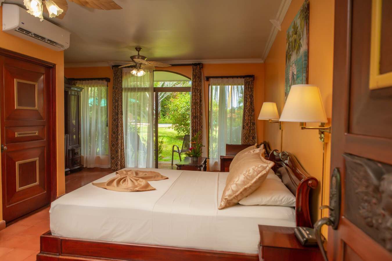 Quarto de hotel com uma cama grande, duas mesas de cabeceira, um ventilador de teto e vista para o exterior.