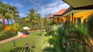 A tropical garden with palm trees, a patio with outdoor furniture, and a building with a red-tiled roof.