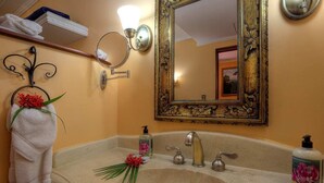 A bathroom with a beige sink, two faucets, a mirror with a decorative frame, a towel rack with a towel and flowers, and a shelf with folded towels.