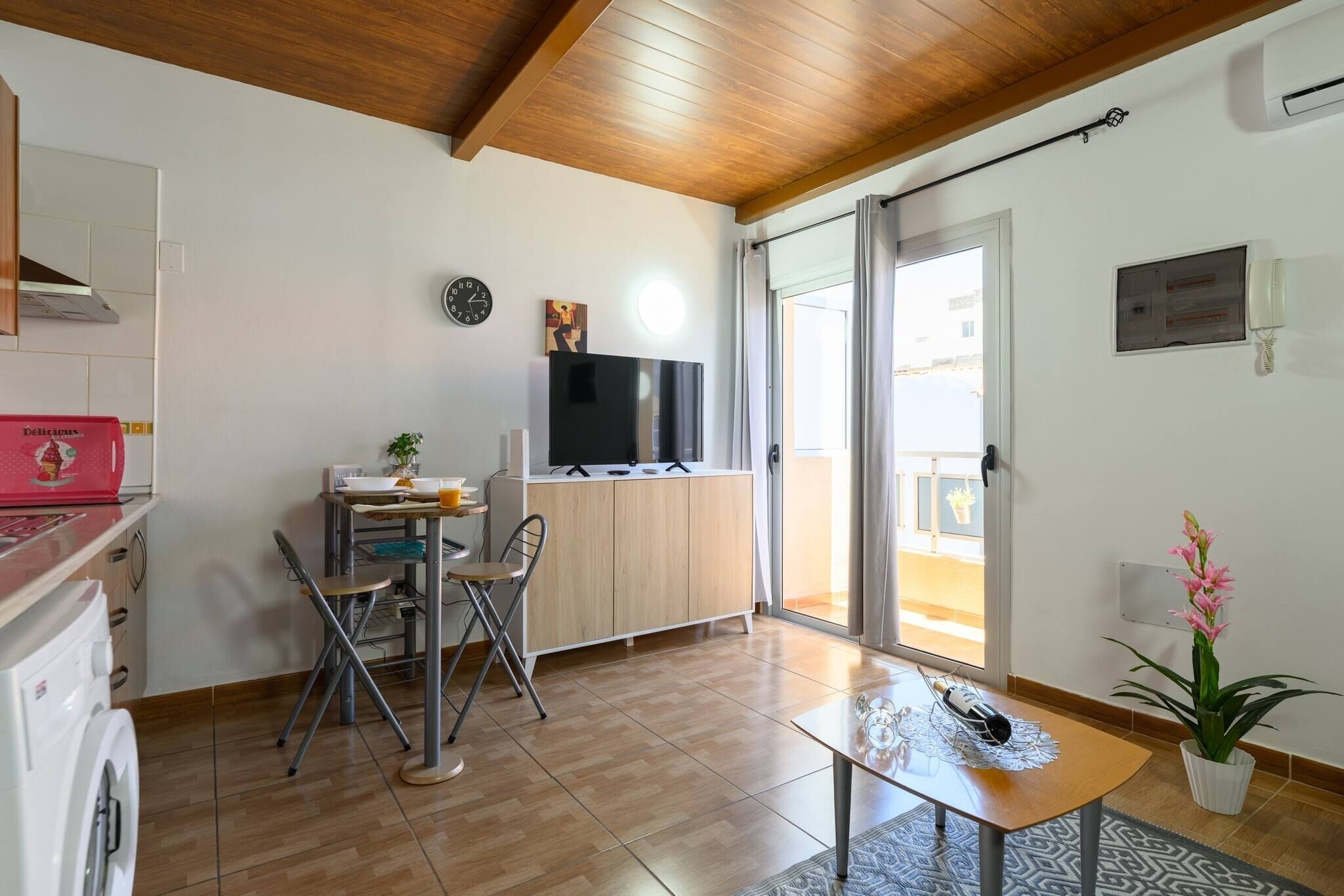 A modern kitchen with a wooden ceiling, a dining area with a table and chairs, a TV, and a sliding glass door leading to a balcony.