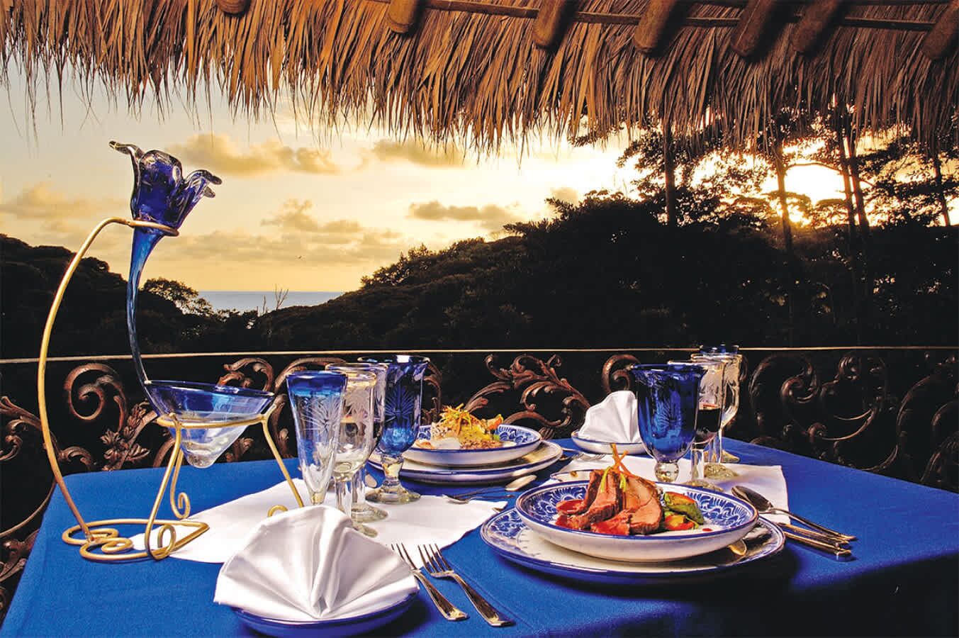 A table set for a meal with a view of the sunset, featuring blue glassware and plates with food.