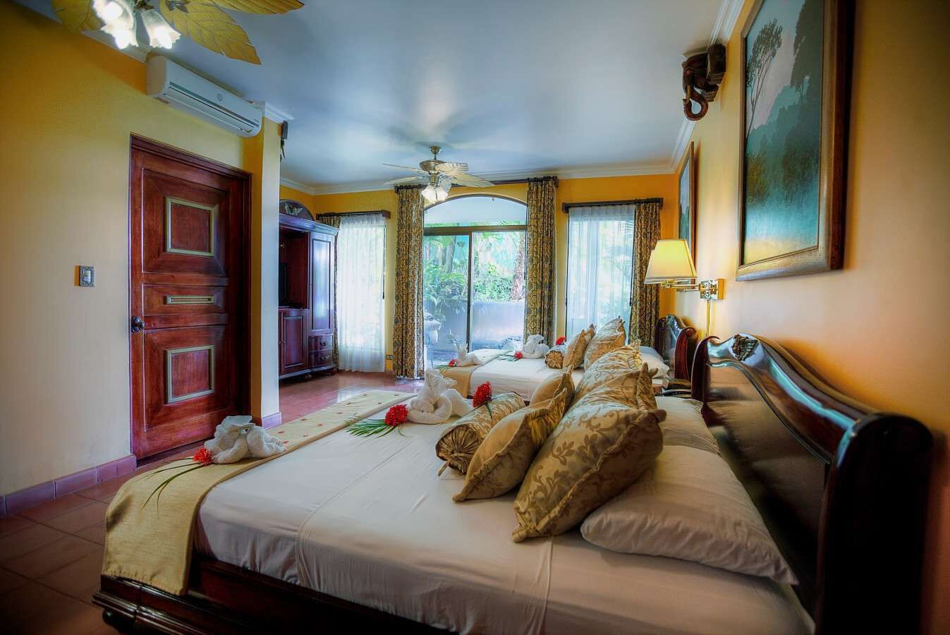 A hotel room with a large bed, a wooden wardrobe, a ceiling fan, and a view of the outdoors.
