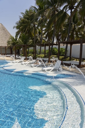 Une piscine d’un bleu éclatant, avec des chaises longues blanches et, au fond, une structure surmontée d’un toit de chaume.